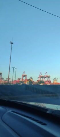 The video shows a view of a cargo port from inside a moving vehicle. The sky is clear and blue. Several large, red cranes are visible in the distance, as well as stacks of shipping containers of various colors. Signs indicating "EXOLGAN CONTAINER TERMINAL" and "INGRESO PARTICULARES" and "SALIDA CAMIONES" can be seen. The foreground shows part of the car's dashboard and exterior. A few lampposts and streetlights are also visible in the background.