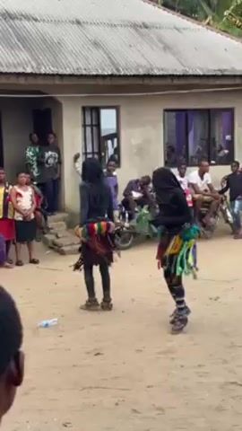 Two individuals dressed in what appear to be masquerade costumes are performing a dance in a sandy courtyard. They are wearing black hooded outfits and colorful skirts, with leg coverings and shoes. The background features a single-story building with two windows. A group of people are observing the performance, some standing on the steps leading to the building. There is also a motorcycle parked nearby. The environment seems to be an outdoor event or gathering.