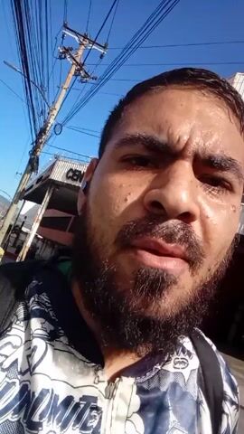 A medium shot video shows a sweaty-faced man speaking to the camera. The man has dark hair and a beard. He is wearing a jacket with a black-and-white pattern and a backpack. He is standing on a city street under a blue sky. There are power lines overhead and buildings in the background. The man encourages viewers to invest.