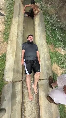 At the 0:00 mark, a young boy slides down a water-filled concrete channel. A man with a beard, wearing a black t-shirt and black shorts with white trim, lies on his back in the channel. Another person, whose lower body is visible wearing white pants and brown shoes, stands to the right of the channel. Two other individuals are partially visible at the top of the channel, one of whom appears to be splashing water. The environment appears to be outdoors with vegetation and dirt surrounding the concrete structure.

By the 0:01 mark, the young boy is still sliding, and the man is in the same position. More individuals are now visible at the top of the channel, with one person in a brown outfit appearing to be pushing or guiding the boy.

At 0:02, the man is still lying down in the channel, and the boy continues to slide. The person in the brown outfit is more clearly visible, and others are observing.

By 0:03, the boy has moved further down the channel, and the man remains in place. The person in the brown outfit is still present.

At 0:04, the boy has reached the bottom of the frame, and the man is still lying in the channel. The other individuals remain in their positions.