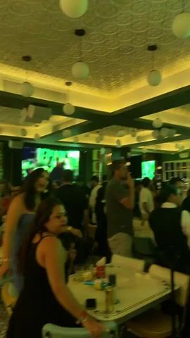 The interior of a busy bar is shown. The room is well-lit with round hanging lights. People are standing and sitting at tables, watching TV screens placed around the bar showing sports. Some people are talking, while others are walking through the room. Tables are decorated with drinks and condiments, and the ceiling features a textured white pattern.