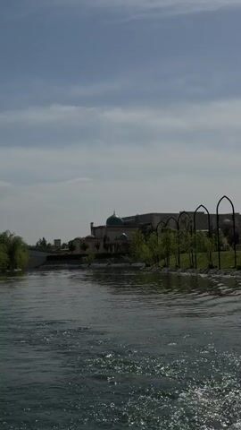 This video captures a scenic view of a river, likely from a moving boat or waterside vantage point. The river occupies the foreground and midground, with gentle ripples visible on its surface.

On the right bank of the river, there's a lush green area with trees and several decorative arched structures. Beyond these, a large building with a prominent green dome stands out against the sky, suggesting a mosque or a similar significant structure. Further in the background, another large building is visible. On the left side of the river, a bridge spans across, and more greenery can be seen.

The sky is partly cloudy, with patches of blue visible. The overall lighting suggests it is daytime, possibly late morning or early afternoon, given the brightness. The architectural style, particularly the green dome and arches, is characteristic of regions like Uzbekistan, aligning with the provided location context of Kuchtumgali. No people or visible text are discernible in the frame.