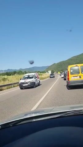 The video shows a view from inside a car driving on a highway during the daytime. The highway has multiple lanes and appears to be experiencing traffic congestion, as several cars are visible in front of the camera. The road is paved and bordered by a grassy shoulder and a metal guardrail on the left. Beyond the shoulder, there are fields of tall grass or crops and a line of trees and hills in the distance. The sky is clear and blue. The car's dashboard is partially visible in the foreground. There are some dark spots on the windshield that obstruct the view slightly. The traffic appears to be moving slowly, as evidenced by the stationary or slow-moving vehicles ahead.