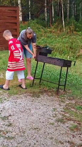 A young boy wearing a red hockey jersey with the number 91 and the name FEDOROV across the back stands to the left of a woman. The woman is wearing a grey t-shirt and denim shorts and is bent over a black barbecue grill. She appears to be tending to something on the grill. The setting appears to be outdoors, possibly a backyard or park, with a wooden fence and trees visible in the background. The ground is covered in gravel and grass.
