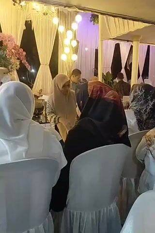 The video shows an indoor gathering, likely a wedding reception or similar event. The scene is decorated with white draping and string lights. In the foreground, two individuals wearing hijabs are seated facing away from the camera. In the background, several people are visible, some appearing to be guests and others possibly staff. One man in a dark shirt and a hat is interacting with another man in a white shirt. There are tables set up with food and decorations. The lighting is dim with warm tones from the string lights.