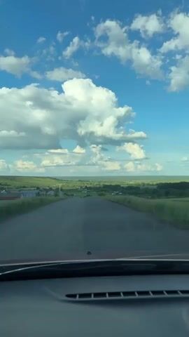 A driver is traveling down a rural road. The landscape is a mix of fields and scattered buildings, stretching into the distance. The sky is a brilliant blue dotted with fluffy white clouds. The view is captured from the interior of a car, focusing on the road ahead.