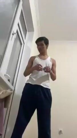 A young man wearing a white tank top and blue sweatpants stands indoors, holding a small white object in his hands and looking down at it. A window with a white frame is to his left, and a beige towel hangs on a ledge below the window. The walls are painted a light beige color. A wall socket is visible near the bottom of the frame.