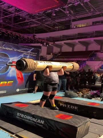 A strongman competitor, wearing shorts, a t-shirt, and knee sleeves, attempts a log press in an indoor arena. He stands between two black elevated platforms, one labeled "PRIDE UNITED" and the other "STRONGMAN International Union of Power Extreme." The competitor lifts a large, rope-wrapped wooden log from the platforms to his shoulders, then drops it back down onto the platforms before clapping his hands.

The arena is lit with purple and pink stage lighting. In the background, empty stadium seating is visible. A large banner with a lightning bolt design and a red fist is prominent. Text on the banner indicates the location: "Sankt-Peterburg, pr. Narodnogo Opolcheniya, 24" and "Stadion «Shtorm»". Other visible text on banners includes "MEGAPARK.WORLD" and "СпортАЙФ" (SportAYF). Several spectators and event staff are standing behind the platforms, watching the event.