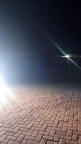 A truck with its headlights on is driving on a brick-paved surface at night. The truck is green and has some lights on its side and back. The headlights of the truck are very bright and cast reflections on the ground. There are also some lights on the truck that are blue and red. The truck is moving from left to right in the frame. The environment is dark and it appears to be an outdoor area.