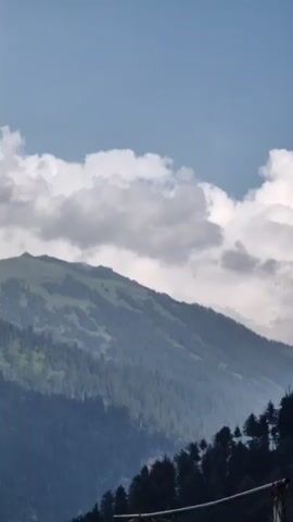 The video displays a time-lapse of a mountain range covered in lush green trees and vegetation. The sky is a clear blue with scattered white clouds that drift and change shape throughout the clip. The perspective is from a slightly elevated viewpoint looking across the valley towards the mountains. The lighting suggests it is daytime, with the sun illuminating the slopes. There are no people or man-made structures visible in the frame.