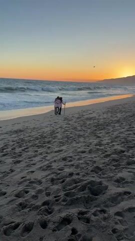 Two people stand on a sandy beach near the water's edge at sunset. The person on the left is wearing a pink jacket and the person on the right is wearing a brown jacket and holding a stick. The sun is setting over the ocean, casting an orange glow across the sky and water. Waves are crashing on the shore. The sand is covered in footprints. A bird is flying in the sky.
