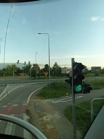 The video captures a view from a vehicle, likely a bus, looking out through the windshield. The vehicle is at an intersection with traffic lights. The traffic lights are green, indicating that it is safe to proceed. There is a crosswalk marked on the road. The intersection is surrounded by grass and trees, and there are signs and streetlights visible. The sky is clear and blue. The video is short and repetitive, with minor variations in the framing of the shot.