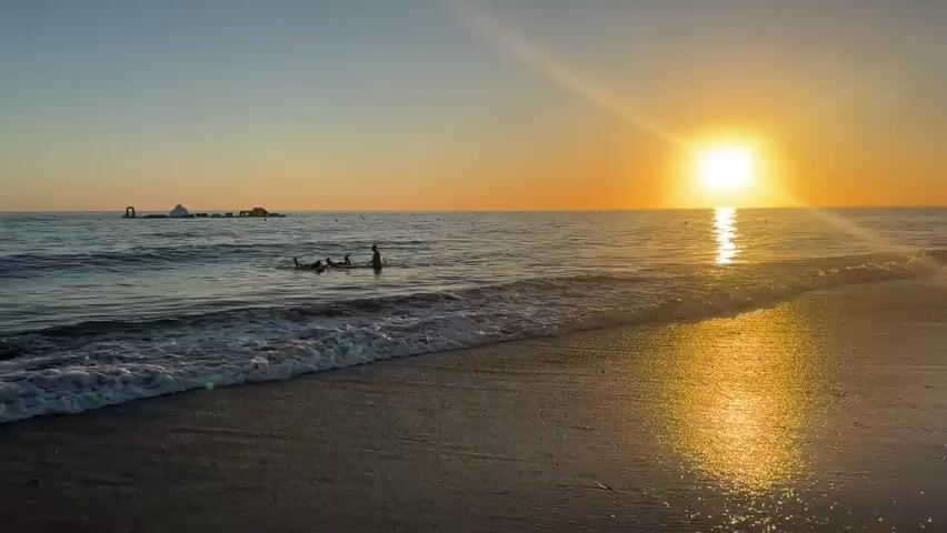 The video shows a beach at sunset. The sun is setting over the ocean, casting a golden reflection on the water. Waves are gently rolling onto the shore. In the water, there are a few people, possibly surfing or playing on paddleboards. In the distance, there appears to be some sort of inflatable water park structure. The sky is a gradient of blue and orange, indicative of a sunset. The overall scene is tranquil and serene.