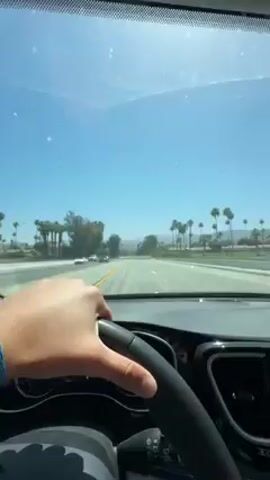 The video captures a first-person perspective from the driver's seat of a car, looking forward through the windshield. A person's right hand, wearing a blue bracelet, is prominently visible on the black steering wheel, positioned at the bottom center of the frame.

The car is in motion, driving along a multi-lane highway or major road. The sky above is a bright, clear blue, indicating a sunny day, likely midday or early afternoon. The bright sunlight creates some glare and reflections on the windshield, which also has a few small specks or dust visible.

The surrounding environment is characterized by numerous tall palm trees lining both sides of the road, extending into the distance. In the far background, faint outlines of arid mountains are visible under the clear sky, consistent with a desert landscape. Other vehicles, including a white sedan and a red car, can be seen in the distance and in adjacent lanes on the road.

The dashboard and a portion of the instrument cluster are visible to the left of the steering wheel, showing some digital gauges and indicators, but no specific text is legible. The overall scene depicts a drive through a warm, open desert region, aligning with the provided context of Palm Desert, United States.