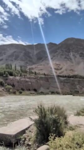 The video captures a scenic view of a wide, fast-flowing river in a mountainous region. The river's water appears somewhat murky, suggesting it might be carrying sediment. On the far bank, steep, eroded cliffs rise, dotted with sparse vegetation and trees. In the distance, large, barren mountains dominate the horizon, their slopes catching the bright sunlight which creates a lens flare effect across the image. The sky above is a vibrant blue with scattered white clouds. In the foreground, the riverbank is rough and unpaved, with some green scrubby bushes and a concrete structure, possibly a small jetty or landing, visible on the left. The overall impression is of a wild, untamed natural landscape.