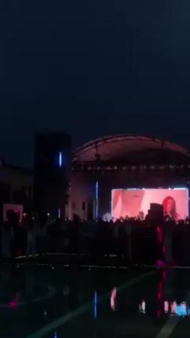 An outdoor concert stage illuminated by blue and pink lights. Silhouetted audience members stand watching a large screen displaying video footage. Puddles reflect the stage lights.