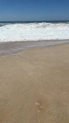 The video shows a sandy beach with the ocean in the background. The sky is clear and blue. Waves are crashing on the shore, creating white foam. The camera is static and positioned at a low angle, looking towards the ocean. The sand appears wet in some areas from the waves. There are no people or other objects visible in the frame. The scene is peaceful and natural.