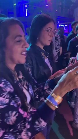 The video shows a crowded event with people holding and using cellphones, presumably recording or taking pictures. The event is lit with blue and purple lights. People are dressed in casual and dressy clothing. Later in the video, people are shown posing on a red carpet in front of a backdrop.