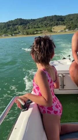 The video captures a bright, sunny day on a calm lake in Golyama Zhelyazna, Bulgaria. The scene is from the perspective of a boat moving through the water, creating a gentle wake.

In the foreground, a young girl in a pink swimsuit adorned with strawberries is seen from the back, looking out at the water. She is holding onto a metal railing on the side of the boat. Her hair is tied up in a bun, and it's slightly windswept.

The lake water is a vibrant green, reflecting the clear blue sky. The sun is high in the sky, casting a bright shimmer on the water's surface. In the distance, rolling green hills covered in trees line the shore, with a few scattered buildings visible on the slopes. The overall atmosphere is one of peace and enjoyment, suggesting a leisurely boat ride on a beautiful summer day. There is no visible text in the media file.
