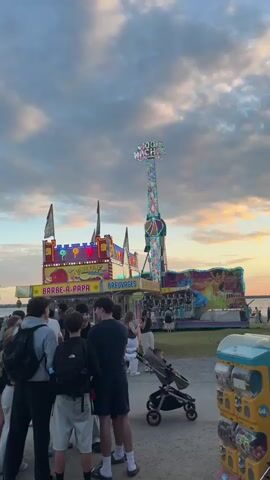 The video shows a tall amusement park ride with flashing lights, labeled "100 NACHT". In the foreground, a group of young people are standing, some facing the ride. To the left, there is a brightly lit booth with signs for "BARBE-A-PAPA" and "BREUVAGES". A few people are visible in the background near the ride and on a sandy area next to a body of water. The sky above is cloudy with hints of sunset colors. A stroller with a child is also visible in the middle ground. The scene appears to be at an outdoor fair or carnival.