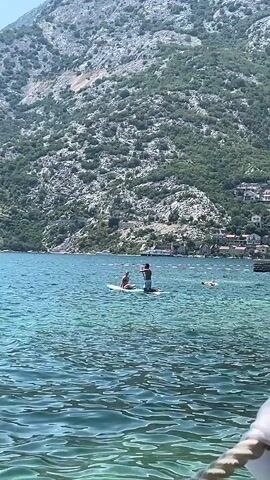 Two people are on a paddleboard on a body of water. The person on the left is sitting down, wearing a pink bikini. The person on the right is kneeling and holding a paddle. The paddleboard is blue. In the background, there is a large, rocky mountain covered in green vegetation. Several buildings are visible on the side of the mountain. The water is clear and blue, with visible ripples. There are other people and boats in the distance. The scene appears to be a sunny day.