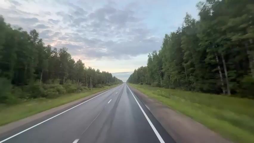 The video shows a POV shot from a vehicle traveling on a wet asphalt road. The road is lined with dense green trees on both sides. The sky is overcast with scattered clouds, and the light suggests it might be dawn or dusk. The road appears to be a highway, with visible lane markings. The motion of the vehicle is smooth and consistent, indicating a steady speed. There are no other vehicles or people visible in the frame. The overall environment is natural and rural.