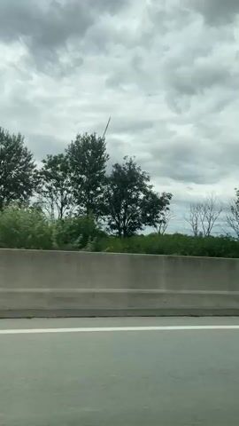 The video shows a side view of a road with a concrete barrier separating it from a natural landscape. A wind turbine is visible in the background, with its blades rotating. Trees and vegetation surround the turbine, and the sky is cloudy. The camera appears to be moving, likely from a vehicle on the road. The lighting suggests it is daytime, but the cloud cover makes it appear overcast. The scene is static, with no human presence or interaction observed.