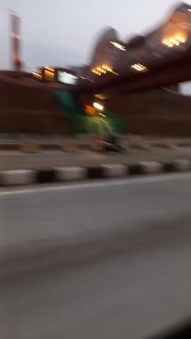 The video shows a view from a moving vehicle of a modern architectural structure. The structure features a prominent, wave-like canopy illuminated with lights, a tall, slender tower, and multiple brick-colored buildings. A sloping, green-lit pathway is visible on the embankment leading up to the structure. The scene appears to be at dusk or dawn, with a pale sky. Concrete barriers line the roadside, and a truck passes by in the background. The camera is at a low angle, looking upwards towards the buildings and the sky.