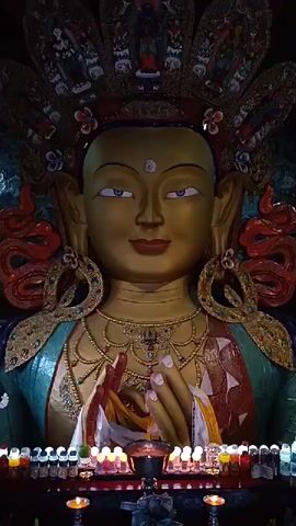 This is a vertical, close-up video of a large, ornate statue, likely of a deity or revered figure, with a golden face and elaborate headdress. The statue has blue eyes, a serene expression, and is adorned with intricate jewelry and colorful fabric. Its hands are held together in front of its chest, wrapped in prayer scarves of white, red, yellow, and blue. In front of the statue, a row of small, lit candles or oil lamps are arranged on a dark surface, possibly an altar. The background is dark and appears to be the interior of a temple or shrine, with decorative elements visible behind the statue's head. The camera slowly zooms in on the statue's face and hands.