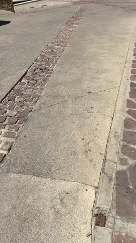 This video shows a street with a stone church in the background. The street is paved with concrete and has a strip of cobblestones running down the middle. There are tables and chairs set up outside a cafe or restaurant to the right of the church. A car is parked on the left side of the street. The sky is blue with some clouds. The video pans up from the street to show the full facade of the church, including its bell tower and a large wooden door.