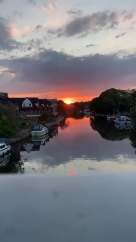 The video shows a sunset over a river. The sky is filled with colorful clouds, and the sun is setting behind a line of trees. The river reflects the colors of the sky, and there are several boats docked along the riverbank. A row of houses is visible on the left side of the river. The overall scene is peaceful and scenic.