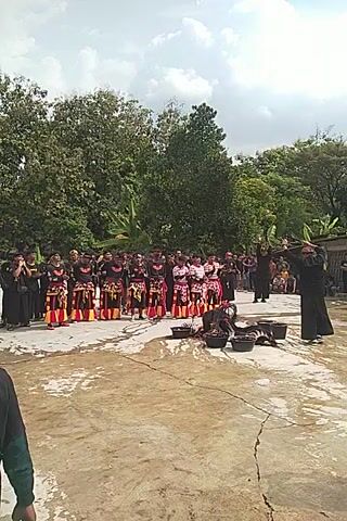 Here is a concise description of the video: 

The video shows a group of people, presumably performers, standing in a row in an outdoor area that appears to be paved, possibly a courtyard. They are wearing traditional outfits with distinct patterns of red, yellow, and black. Some individuals in black attire are standing in front of the performers. The backdrop consists of lush green trees and a cloudy sky. There are also some speakers or musical instruments set up in front of the group. The video is filmed at a slightly tilted angle.