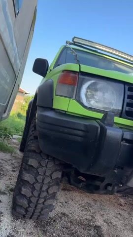 This is a low-angle, close-up shot of the front driver's side of a lime green Mitsubishi Pajero. The vehicle has large, knobby off-road tires and a light bar mounted on the roof. The vehicle is parked on a dirt and gravel surface, with a white structure visible on the left and greenery in the background. The sky is blue. The video appears to be a still frame with no discernible action.