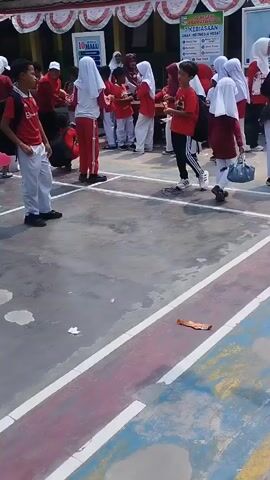 The video opens with a wide shot of an outdoor schoolyard. Two boys in red shirts and white pants face each other, one wearing a backpack, as other students in similar attire mill about in the background. A pink balloon is visible on the ground. The camera pans to the right, showing more students, some wearing hijabs, gathered near a building adorned with red and white banners. Several motorcycles are parked in front of the building. The camera then moves to a sign that reads "SDN KRAMATWATU 2 SEKOLAH RAMAH ANAK (SRA)" and lists NPSN 20604908. Further right, another sign displays "8 DIMENSI PROFIL KELULUSAN SD NEGERI KRAMATWATU 2". The video concludes with a close-up shot of two parked motorcycles in front of a building with wooden chairs stacked inside. The ground is paved with concrete marked with white and red lines, and there are scattered pieces of trash.