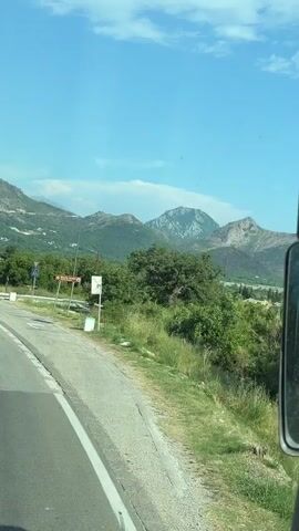 The video captures a scenic drive through a mountainous landscape, likely in Kaluđerac, Montenegro. The camera is positioned as if from a vehicle, showing the road ahead and the surrounding environment. 

The scene is set on a sunny day, with a clear blue sky dotted with a few wispy clouds. Rolling hills and majestic mountains dominate the background, their slopes covered in lush green vegetation. In the mid-ground, there are scattered houses and trees, suggesting a rural or semi-rural setting. The road itself is paved, with white dashed lines indicating lanes. There are several road signs visible. One sign, with a red border and a number 50 inside a circle, indicates a speed limit. Another sign with a red circle and a diagonal bar across it suggests a prohibition, likely related to traffic. There is also a sign with an arrow pointing to the right and some text in a foreign language, possibly indicating directions to a town or attraction.

The activity taking place is a drive through this picturesque region. The camera's perspective, moving along the road, conveys a sense of journey and exploration. The overall mood is peaceful and serene, enhanced by the natural beauty of the landscape and the pleasant weather. No people are directly visible in the footage, but the presence of houses and the road suggests human habitation and activity in the area. The focus is primarily on the visual experience of traveling through this scenic location.