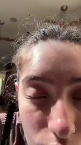 A close-up video shows a person applying black mascara to their upper eyelashes. The person has fair skin, light brown eyebrows, and light-colored eyes. The person is wearing a brown shirt. The background is out of focus but appears to be indoors.