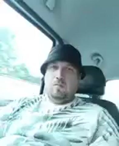 The video shows a man sitting in a vehicle, likely a car, with his head tilted slightly upwards and to the side. He is wearing a black bucket hat and a t-shirt with a light-colored, possibly white, palm leaf pattern. The man has a beard and appears to be in his late 30s or 40s. His expression is neutral or perhaps slightly contemplative.

The setting suggests an outdoor environment, possibly a road trip, as trees and foliage are visible through the vehicle's window on the left side. The sky appears overcast or hazy, indicating a cloudy day or a bright, diffused light. The interior of the car is visible, including the headrest of a seat behind the man and part of the roof lining.

The activity is static, with the man simply seated in the vehicle. There is no explicit action occurring, and the focus is on the man's presence within the car and the surrounding environment. The video appears to be a short, unedited clip, possibly a selfie or a quick recording of the man during transit. No text is visible in the media file. The time of day is difficult to ascertain precisely due to the overcast conditions, but it is clearly daytime. The location context of Říčany, Czechia, suggests the scenery might be typical of the Czech countryside or suburban areas.