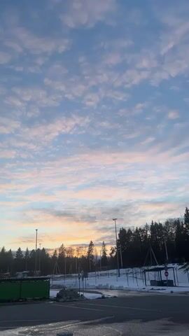 The video presents a serene winter landscape, likely during sunrise or sunset, in Lupanovo, Russia. The sky is a prominent feature, adorned with numerous scattered clouds displaying delicate hues of pink, orange, and light blue against a pale blue backdrop.

In the mid-ground, a dark line of evergreen trees, possibly a forest edge, extends across the horizon, with a soft, warm glow visible just above them, indicating the sun's position. Tall streetlights punctuate the scene, suggesting an artificial lighting infrastructure.

The foreground consists of an asphalt road or parking area, partially covered in patches of snow and ice. To the left, a large, dark green container is visible. To the right, a bus stop shelter, also partially covered in snow, features a red "STOP" sign. Other traffic signs are present but not clearly legible. Further in the distance, behind the trees, glimpses of buildings or vehicles can be seen.

The video is largely static, capturing the peaceful scene as the camera subtly zooms out over the first few seconds. A male voice speaking in Russian is heard, saying "Блядь, вы ж все ждали, бля, этот момент, нах." (informal/expletive-laden, translates to "Damn, you all were waiting for this moment, huh."), contrasting with the calm visual. The overall impression is one of a quiet, picturesque winter morning or evening.