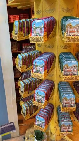 The camera pans slowly to the right, revealing a spinning, decorative display in a shop. The display has three tiers of product, canned fish, each tin with a different city depicted: Aveiro, Coimbra, and Lisbon. The shop has wooden floors. A tall shelf behind the display is covered with hundreds of cans. A small potted succulent is visible on the bottom level.
