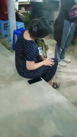 In a casual indoor or semi-outdoor setting in Kebonsari, Indonesia, a woman with dark hair tied back is seen sitting on a concrete floor. She is wearing a navy blue dress with white polka dots and sandals. A child, partially visible in a camouflage-patterned top, is sitting on her lap or very close to her. A black smartphone is placed on the floor next to her. In the background, other people are present: one person is standing wearing gray pants and white sneakers, and another person in a red shirt is seated on a blue plastic stool. The overall scene appears to be a relaxed social gathering.