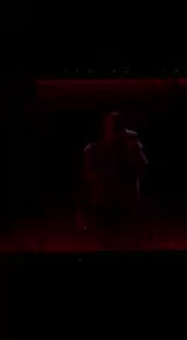 A live performance is captured in a dark setting, likely a concert stage at night in Indio, United States. A lone figure, presumed to be a singer or performer, is seen in silhouette against a backdrop of intense red stage lighting. The performer holds a microphone and moves dynamically across the stage, which is mostly obscured by darkness, with only faint red glows indicating its boundaries. The strong red lighting from below and behind creates a dramatic and atmospheric visual, highlighting the performer's form while keeping the details of their appearance indistinct. The video features music playing, with a clear vocal track, suggesting an emotional or energetic musical piece. The camera occasionally shifts focus or cuts, but the overall impression is of a theatrical and somewhat mysterious live act, emphasizing the performer's presence through light and shadow.