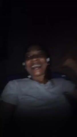 A woman smiles and laughs into the camera. She is wearing a gray t-shirt and has white earbuds in her ears. The background is dark and appears to be indoors. The lighting is dim, and the woman's face is illuminated.