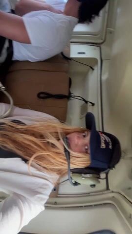 A woman with long, blonde hair wearing a dark blue baseball cap and a headset is in the passenger seat of a helicopter. She is looking directly at the camera. Behind her, a person in a white t-shirt is also seated, buckled in with a seatbelt and wearing a headset. A gold purse sits on the seat beside the woman. The interior of the helicopter is visible, with light beige walls and brown leather seats. The helicopter appears to be in flight, with a view of the landscape visible through the windows.