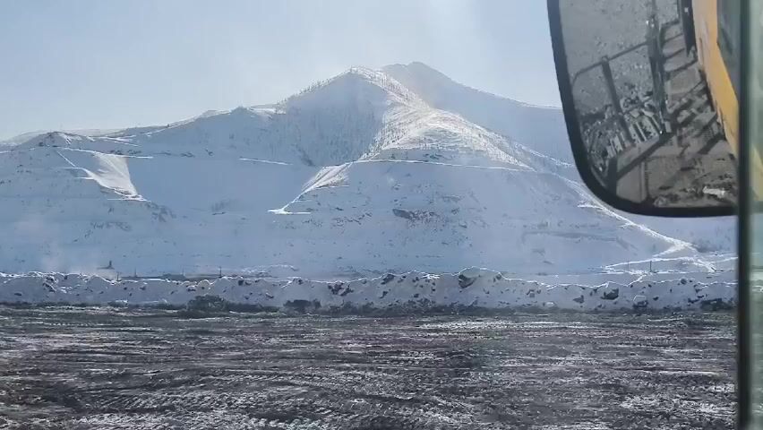 The video shows a snowy landscape as seen from inside a vehicle. A large mountain covered in snow is the central focus, with visible terraced layers suggesting mining activity. The ground in the foreground is a dark, textured surface with patches of snow. A side mirror of the vehicle partially obstructs the view on the right side of the frame. The sky is bright and clear, indicating a sunny day.
