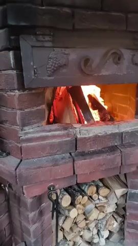 This video shows a close-up shot of a brick fireplace with a fire burning inside. The logs in the fireplace are burning brightly, and the flames are visible. Below the fireplace, there is a stack of firewood. The fireplace has an ornate metal decoration above the opening, featuring grape clusters and swirling patterns. The video pans slightly to the right, revealing more of the brickwork and a glimpse of something dark and possibly fabric to the far right. The overall impression is one of warmth and coziness.