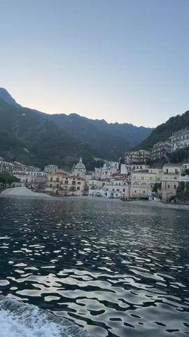 The video shows a shoreline town in Italy, likely on the Amalfi Coast, from the perspective of a boat on the water.  The town is nestled between a beach and steep, forested hills. Buildings are closely packed, many painted in light colors. A notable building is a domed church, and a stone tower sits on a rocky promontory at the end of the town. The water is dark and rippled, reflecting the sky. The sky is a clear, pale blue, suggesting early morning or late afternoon. The boat is moving, creating a wake.