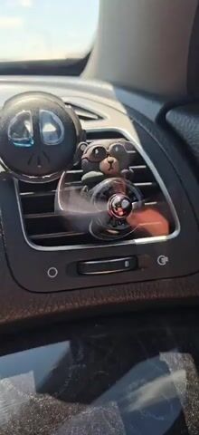 A black car air vent is visible with a rotating fan attached. The fan has a small black bear figure wearing sunglasses sitting on top of it, appearing to ride the fan. To the left of the fan, a circular black object with a peace symbol design is also attached to the vent. The fan is in motion, creating a blur effect. The background shows the interior of a car, with a dashboard and possibly a window visible.