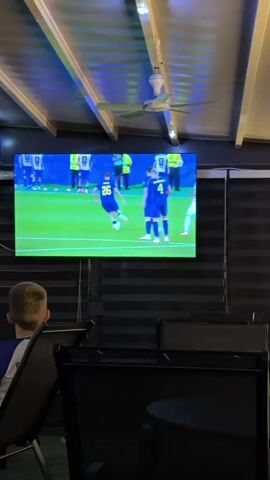 A wide, flat-screen television is mounted on a dark, horizontally striped wall. The television displays a soccer match with players in blue and white uniforms on a green field. The television is brightly lit, and the ambient lighting in the room is dim and colored blue and white. A white ceiling fan with three blades is visible in the upper right corner of the image. In the foreground, a young boy with blond hair sits facing away from the camera, looking at the television. He is wearing a blue and white shirt. The boy is seated in a dark chair. Several other dark chairs and tables are visible in the dim foreground.
