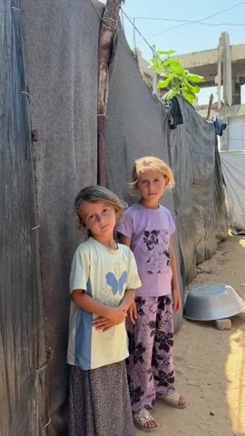 Two young girls, one with blonde hair and the other with brown hair, stand in what appears to be a temporary shelter or camp. The girl on the left wears a light-colored t-shirt with a butterfly graphic and a long, patterned skirt. The girl on the right wears a light purple t-shirt and patterned pants. They are standing on sandy ground next to tents made of dark fabric. In the background, a partially constructed building with visible rebar suggests ongoing development or damage. The sky is clear and blue.