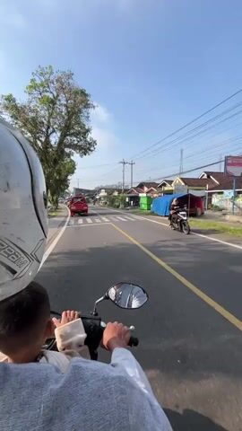 The video, shot from the perspective of the passenger on a motor scooter, shows a view of a road in a rural or suburban area on a clear, sunny day. The road is asphalt with a yellow center line and a crosswalk visible in the distance. Trees line the left side of the road, and houses and utility poles are on the right. A red truck is traveling ahead of the scooter.

The scooter is being driven by an adult, visible only by their hands on the handlebars and the reflection of their shirt in the scooter's mirror. A child wearing a helmet is seated in front of the driver, their back to the camera. Other scooters pass by on the left side of the road. The sky is blue with scattered clouds.